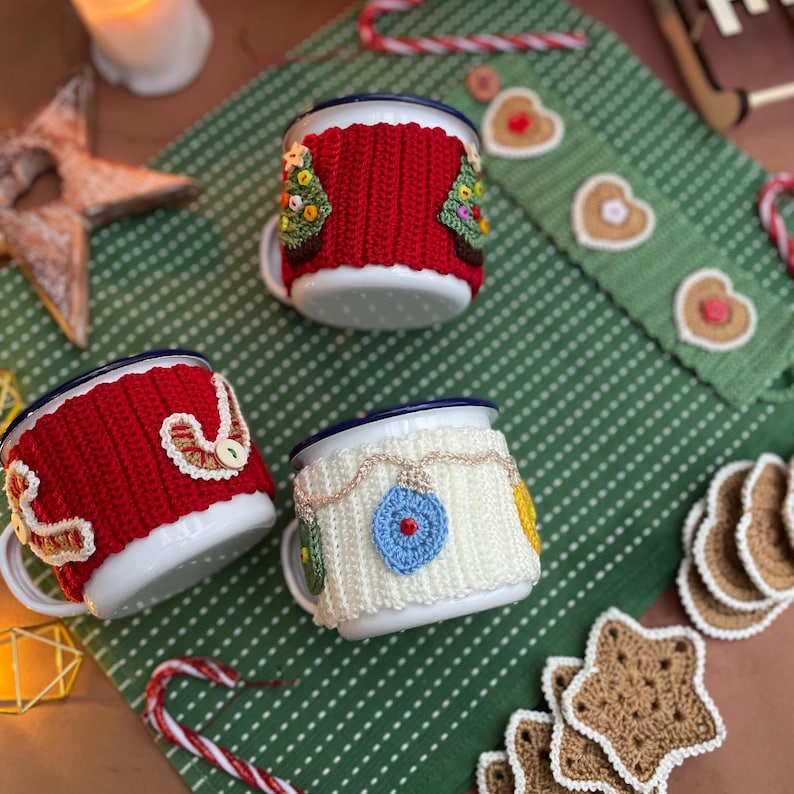May include: Three white enamel mugs with blue rims, each adorned with a unique, colorful, crocheted cozy. One mug features a red cozy with a Christmas tree design, another with a white cozy and ornament design, and the third with a red cozy and button design.