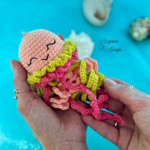 May include: A crocheted jellyfish toy with a pink body, green and pink tentacles, and a smiling face. The toy is being held in a person's hands.