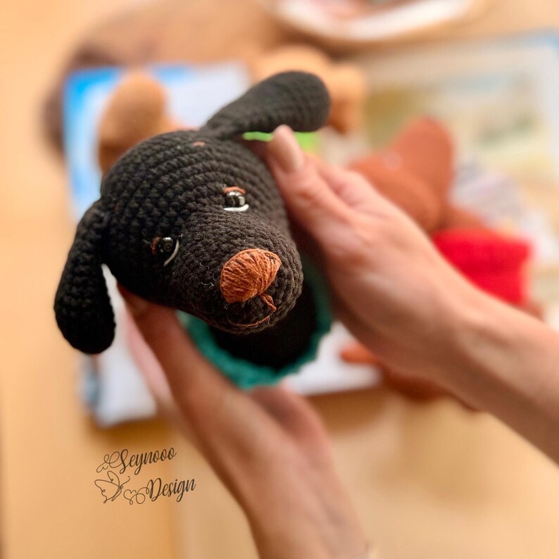 May include: A handmade crochet dog with a black body, brown nose, and small, detailed eyes. The dog's head is held up, showcasing its features. The image includes the text "Seynooo Design" in the bottom left corner, suggesting a craft item.