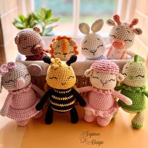 Könnte beinhalten: Eine Sammlung handgefertigter Häkeltierpuppen, darunter ein Bär, ein Löwe, ein Hase, eine Giraffe, ein Schwein, eine Biene, ein Schaf und eine grüne Kreatur. Jede Puppe hat geschlossene Augen und trägt verschiedene Outfits. Die Puppen befinden sich in einem Weidenkorb.