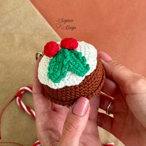 May include: A crocheted Christmas pudding ornament with green holly leaves and red berries. The ornament is brown, white, and green.