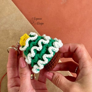 May include: A green and white crocheted Christmas tree keychain with a yellow star on top. The keychain is attached to a silver key ring.