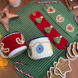 May include: Two white enamel mugs with blue rims, adorned with red and white knitted cozies. One cozy features a decorative design, the other has a blue ornament. Christmas-themed knitted mug cozies and coasters are also visible.