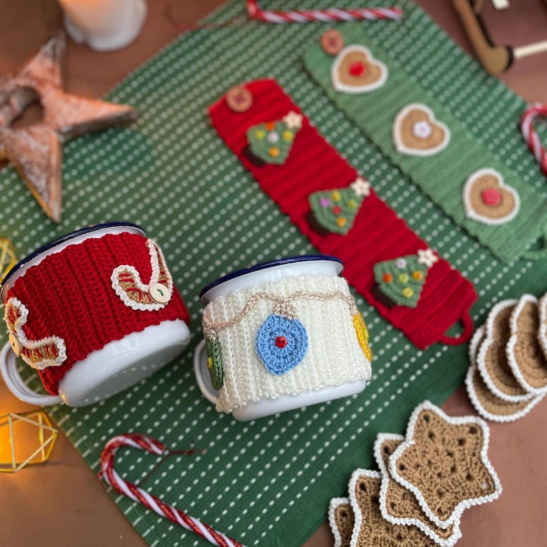 May include: Two white enamel mugs with blue rims, adorned with red and white knitted cozies. One cozy features a decorative design, the other has a blue ornament. Christmas-themed knitted mug cozies and coasters are also visible.