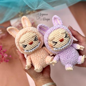 May include: Two handmade crochet bunny dolls in peach and lavender. Each doll has a smiling face with closed eyes, a red nose, and a white-stitched grin. The dolls are wearing hooded outfits. The text "Seyroco Design" is visible in the background.
