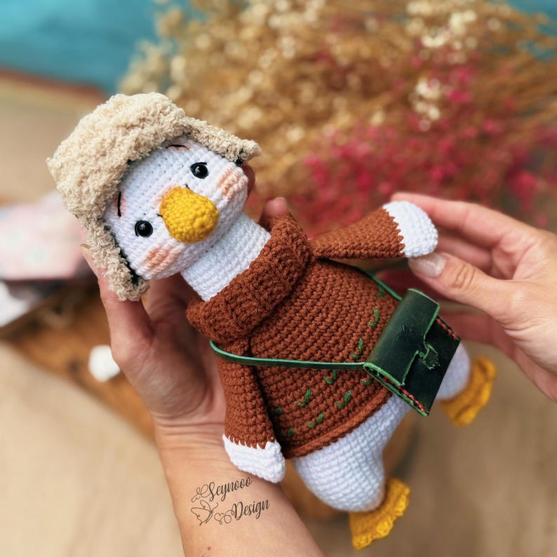 May include: A handmade crochet duck doll wearing a brown sweater, a beige hat, and a green leather bag. The duck has a white body, a yellow beak and feet, and black eyes. The doll is held in a person's hands.
