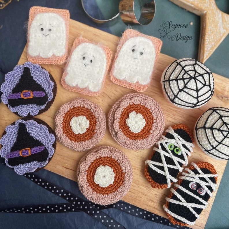 May include: A collection of crocheted Halloween-themed decorations, including ghosts, pumpkins, a spiderweb, and mummies. The decorations are arranged on a wooden surface with a black and white polka dot ribbon.