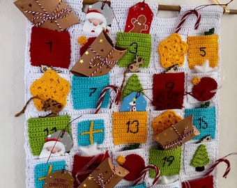 May include: A handmade Christmas advent calendar with 25 numbered pockets in various colors. Each pocket contains a small gift or decoration, including star-shaped ornaments, gift boxes, and candy canes. The calendar is crocheted with a white background.