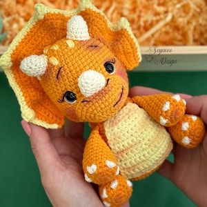 Peut inclure: Un jouet en crochet en forme de tricératops, de couleur orange et jaune, avec des accents blancs. Le jouet a une expression amicale et tient ses bras tendus.
