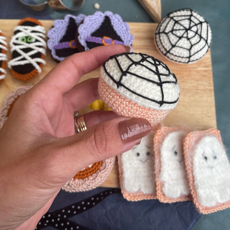 May include: A collection of crocheted Halloween-themed treats, including a spiderweb, a mummy, a witch's hat, and ghosts. The treats are made with a variety of colors, including white, black, orange, and purple.
