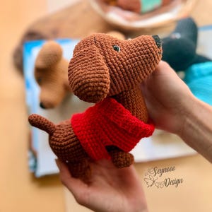 May include: A crocheted brown dachshund dog wearing a red sweater. The dog has a black nose and small black eyes. The dog is held in a person's hands. The background includes other crocheted dogs.