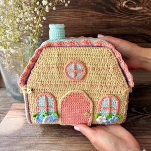 May include: A crocheted house-shaped container in pale yellow with a pink arched door, light blue windows, and a matching chimney. The roof is trimmed with a scalloped pink border, and small flower-shaped buttons accent the sides and door handle.