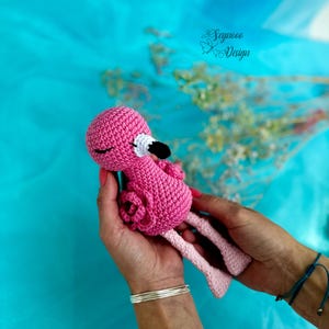 May include: A pink crocheted flamingo toy with black eyes and a white beak. The flamingo is being held in two hands.
