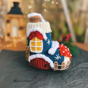 Può includere: Una calza all'uncinetto a forma di casa con base blu, porta rossa e finestra gialla. La parte superiore presenta un risvolto color crema con un bordo bianco soffice che ricorda la neve. Un tetto rosso a pois bianchi copre la punta.