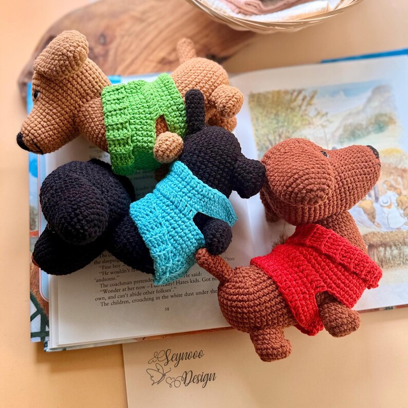 May include: Crocheted dachshund dog toys in various colors, including brown, black, and red, each wearing a tiny sweater. The toys are arranged on an open book with text and illustrations.