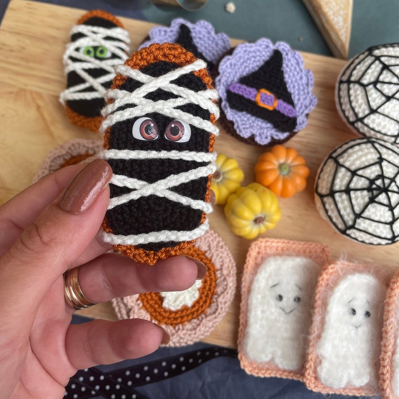 May include: A collection of crocheted Halloween-themed decorations, including a mummy, a witch's hat, a ghost, and a spider web. The decorations are made with black, white, orange, and purple yarn.