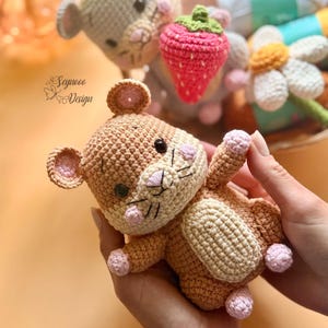 May include: A crocheted brown mouse toy with pink accents on its ears, cheeks, and paws. The mouse has black eyes and a stitched nose and mouth. Other crocheted toys, including a strawberry and a daisy, are visible in the background. The image includes the text "Seynooo Design".