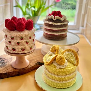 Peut inclure: Trois gâteaux crochetés sont présentés. L'un est blanc avec des fraises rouges, un autre est marron avec des rayures rouges et blanches, et le troisième est jaune avec des tranches de citron. Les gâteaux sont sur des présentoirs et une planche en bois.