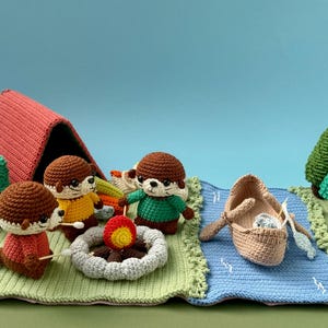 May include: A crochet playset featuring three brown otters, a red tent, a campfire with a yellow and red flame, a tan boat with oars, and several green trees. The scene is set on a green and blue crocheted mat representing land and water.