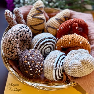 Op de afbeelding: Een verzameling gehaakte voedingsmiddelen in een rieten mand. De items omvatten een donut, een pretzel, een croissant, een spiraalvormig brood en een chocolade-peer.