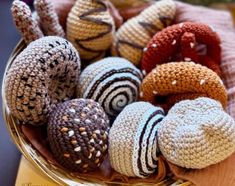 Op de afbeelding: Een verzameling gehaakte voedingsmiddelen in een rieten mand. De items omvatten een donut, een pretzel, een croissant, een spiraalvormig brood en een chocolade-peer.