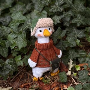 May include: A crocheted goose doll wearing a brown sweater, a green and beige hat, and a small green bag. The goose has a yellow beak and feet, and is standing in front of green foliage.