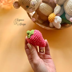 May include: A handmade, crocheted strawberry with a green stem and pink body with white speckles. The strawberry is held in a hand, with other crocheted toys in the background. The image includes the text "Seynooo Design".