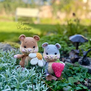 May include: Two crocheted hamster dolls. One is brown and tan, holding a white and yellow flower. The other is blue and tan, holding a pink strawberry. The dolls are set in a garden with greenery and a blurred background.