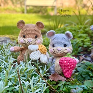 May include: Two crocheted mouse dolls, one brown and one gray, are featured in a garden setting. The brown mouse holds a white and yellow flower, while the gray mouse holds a pink strawberry. The dolls have pink cheeks and are surrounded by greenery.