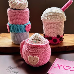 Könnte beinhalten: Drei gehäkelte Amigurumi-Spielzeuge in Form eines Cupcakes, eines Boba-Tees und einer Kaffeetasse. Der Cupcake ist rosa und weiß mit einem blau-weißen Streifen um die Mitte. Der Boba-Tee ist rosa mit braunen Boba-Perlen und einem weißen Deckel. Die Kaffeetasse ist rosa mit einem weißen Herz auf der Seite. Alle drei Spielzeuge haben ein herzförmiges Design auf der Seite.