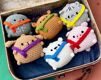May include: Six crocheted animal plush toys are displayed in a vintage suitcase. The toys include a grey owl, a brown reindeer, a grey hippo, a pink pig, a brown dog, and a white bear, each with a colourful scarf. The suitcase is brown with a leather handle.