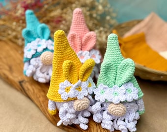 May include: Four colorful crochet gnomes with white beards and flower accents. The gnomes have pointed hats in blue, pink, yellow, and green. One yellow gnome has bunny ears. The gnomes are displayed on a wooden board.