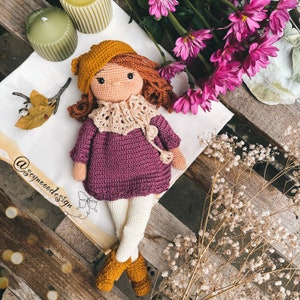 May include: A crocheted doll with brown hair, wearing a purple dress, a beige scarf, and a brown hat. The doll is sitting on a wooden surface with dried flowers and a piece of paper with the text "@seynoodedesign".