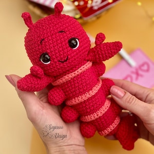 Könnte beinhalten: Ein gehäkelter roter Hummer mit rosa Streifen und großen schwarzen Augen. Der Hummer lächelt und hat einen freundlichen Ausdruck.