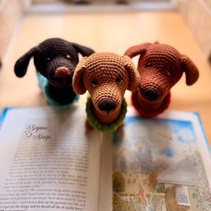 May include: Three handmade crochet dachshund dog figurines in black, brown, and tan, each wearing a different colored collar. The dogs are positioned on an open book. The image includes the text "Seywoo Design".