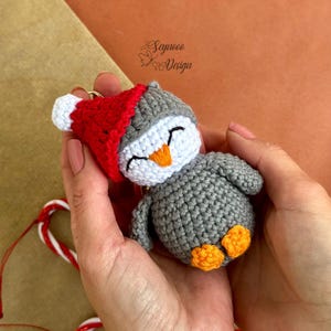 May include: A grey crocheted penguin wearing a red Santa hat with white trim. The penguin has a white face with an orange beak and black eyes. The penguin is holding a key chain.
