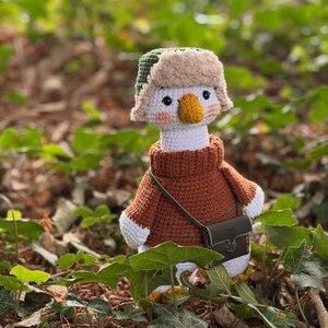 May include: A handmade crochet goose wearing a brown sweater, a green and tan hat, and a small green bag. The goose has a white body, a yellow beak, and is set against a blurred background of green foliage.