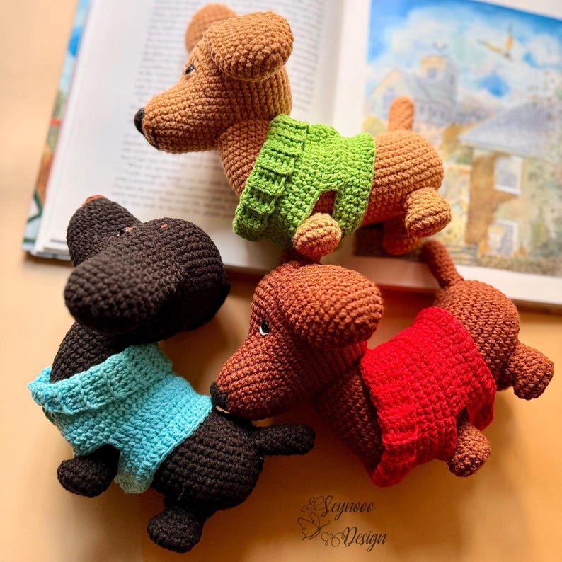 May include: Four crocheted dachshund dog toys in various colors: brown, black, and two shades of brown. Each dog wears a sweater in green, blue, and red. The toys are arranged on a light orange surface with a book in the background.