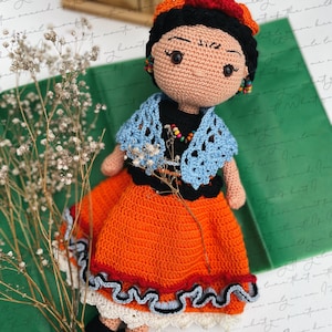 May include: Crocheted doll with black hair, a red flower crown, and a blue and orange dress. The doll is wearing a black shawl with a blue lace trim. The doll is standing on a green surface with white flowers.