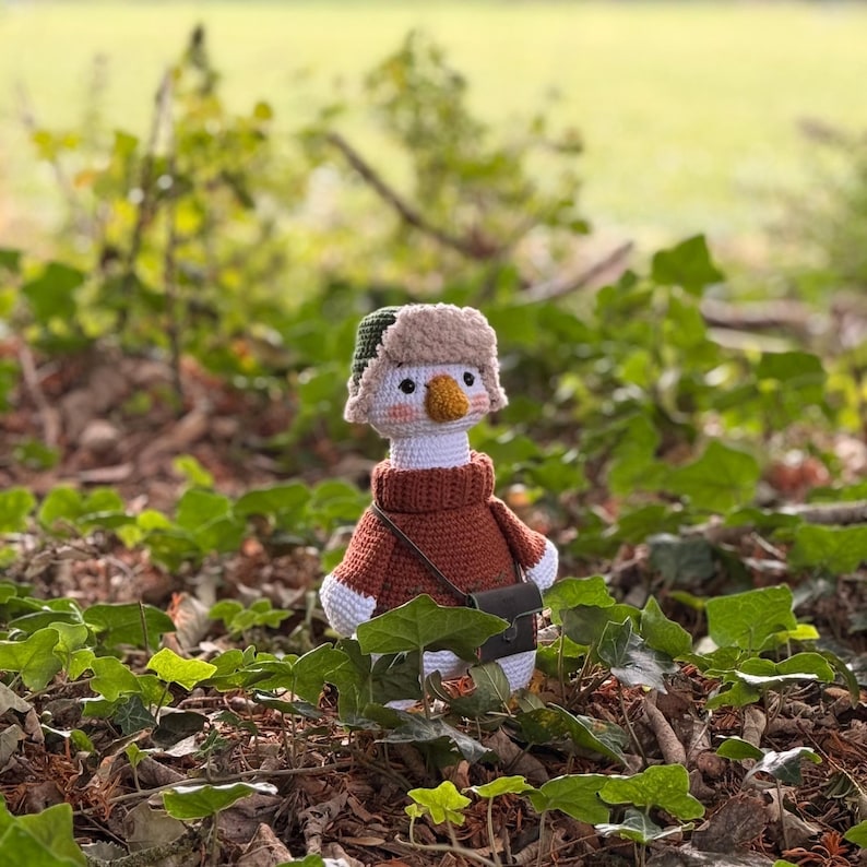 May include: A handmade crochet goose wearing a brown sweater, a fur-lined hat, and a small black bag. The goose has a white body, a yellow beak, and is set against a backdrop of green foliage.