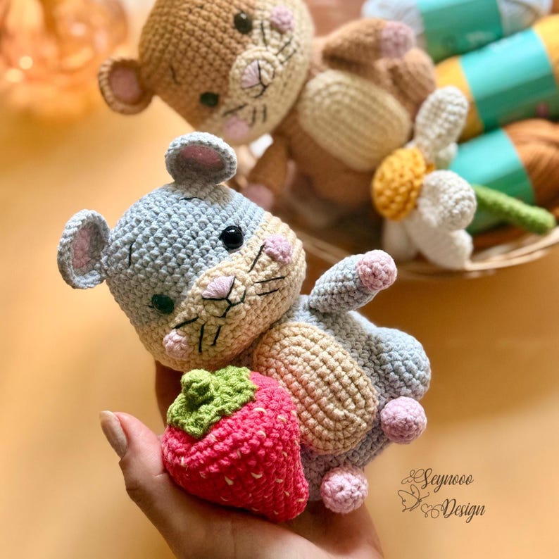 May include: Crocheted mouse toy in gray, beige, and pink, holding a red strawberry with a green top. Another crocheted mouse toy is in the background. The toys are handmade and have black eyes and pink accents.
