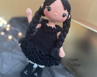 Könnte beinhalten: Handgemachte Häkelpuppe mit schwarzen Zöpfen, trägt ein schwarzes Kleid und schwarze Schuhe. Die Puppe hat ein hellrosa Gesicht und Arme. Das Kleid der Puppe hat einen gerüschten Ausschnitt und Rock. Es ist ein handgefertigter Artikel.