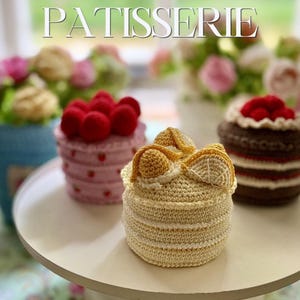以下が含まれることがあります： 上部にPATISSERIEの文字がある3つのクロッシェ編みのケーキ。手前のケーキはクリーム色で黄色い柑橘系のスライスがのっています。左側は赤いベリーがのったピンクのケーキ、右側は赤いベリーと白いクリームがのった茶色のケーキです。
