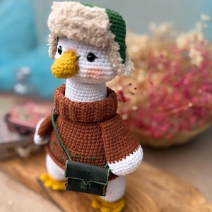 May include: A handmade crochet duck doll wearing a brown sweater, a green and beige hat, and a small green purse. The duck has a yellow beak and feet, and white body. The doll is standing on a wooden surface.
