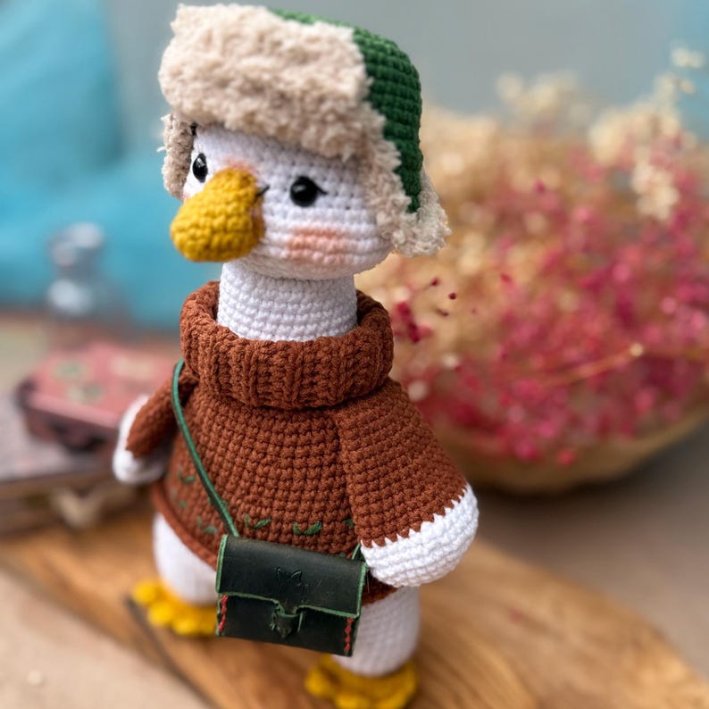 May include: A handmade crochet duck doll wearing a brown sweater, a green and beige hat, and a small green purse. The duck has a yellow beak and feet, and white body. The doll is standing on a wooden surface.