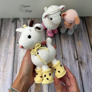 May include: Three handmade crochet amigurumi toys including a white goat with brown horns and yellow boots, a white sheep with pink boots, and a pink pig with grey shoes. The goat features a yellow ribbon tied around its neck and black eyes.