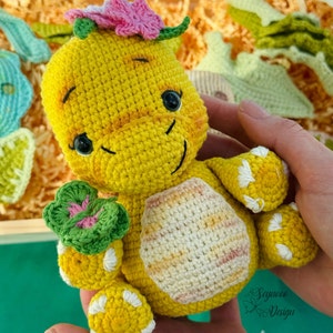May include: A crocheted yellow dinosaur toy with a pink flower on its head and a green clover in its hand. The dinosaur has a white and yellow striped pattern on its back.