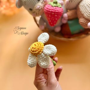 May include: A handmade crochet daisy flower with white petals and a yellow center. The flower is held in a hand, with other crochet toys in the background. The text "Seynooo Design" is visible.