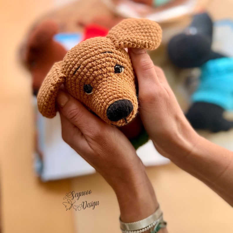 May include: A crocheted brown dog with a black nose and small black eyes. The dog has floppy ears and is held in a person's hands. The dog is a handmade item.