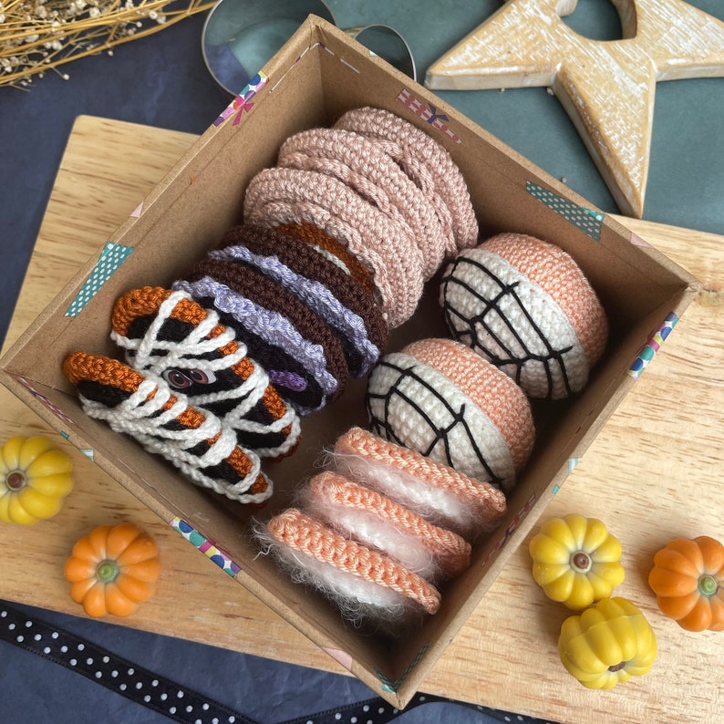 May include: A box filled with crocheted food items, including a stack of pink donuts, a stack of brown and purple donuts, a spiderweb-patterned orange and white donut, a stack of pink and white donuts, and a spiderweb-patterned orange and white donut.
